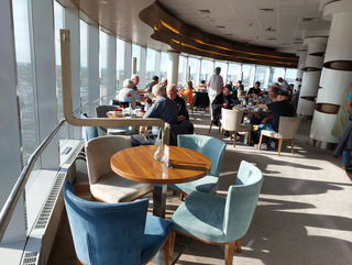 Helles, modernes Restaurant in einem Hochhaus mit Panoramablick auf die Stadt. Gäste genießen Kuchen und Getränke im Sonnenlicht.