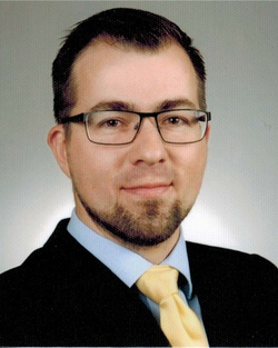 Porträt eines Mannes mit kurzen dunklen Haaren, Bart, Brille, dunklem Anzug, hellblauem Hemd und gelber Krawatte.