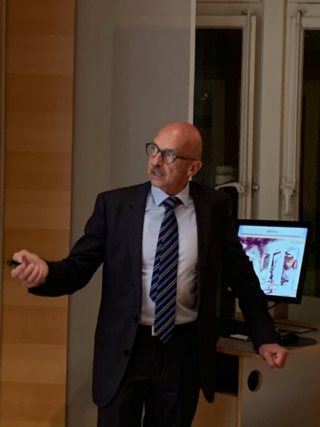 Ein kahler Mann in Anzug und Krawatte mit Brille spricht während einer Präsentation mit einem Bildschirm im Hintergrund.