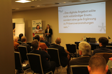 Zwei Frauen präsentieren vor einem Publikum bei einer Konferenz. Eine Leinwand zeigt eine Präsentation über digitale Angebote.