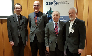 Vier lächelnde Männer in Anzügen stehen vor einem Banner der GSP (Gesellschaft für Sicherheitspolitik e.V.).
