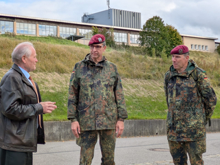 Drei Männer, ein Zivilist und zwei deutsche Soldaten in Tarnuniform mit roten Baretten, unterhalten sich draußen vor Gebäuden.