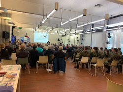 Gemischtes Publikum, darunter Soldaten in Tarnuniformen und Zivilisten, verfolgt eine Präsentation in einem großen Saal.