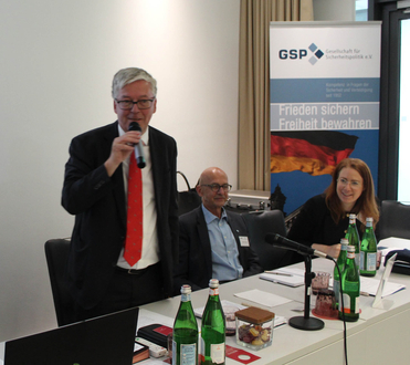 Ein Mann spricht ins Mikrofon, flankiert von zwei Personen an einem Tisch, vor einem "GSP"-Banner.
