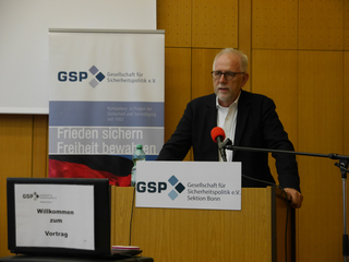 Ein Mann spricht an einem Pult vor GSP-Bannern. Ein Schild auf dem Pult sagt 'Willkommen zum Vortrag'.