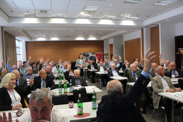 Teilnehmer einer Konferenz oder Versammlung heben die Hände, einige sitzen an Tischen mit Wasserflaschen, eine Frau steht vorne.