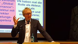 Mann in Anzug und Brille spricht am Rednerpult, gestikulierend. Im Hintergrund Folien zu Global Governance und UN-Rolle.
