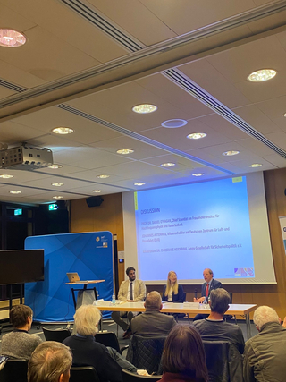 Podiumsdiskussion mit drei Sprechern und Publikum in einem Konferenzraum. Eine Leinwand zeigt 'DISKUSSION' und Namen.