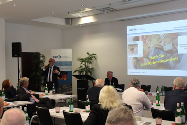 Ein Sprecher präsentiert auf einer Konferenz vor Publikum; im Hintergrund GSP-Banner und Euro-Banknoten auf der Leinwand.