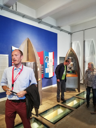 Ein Museumsführer spricht, während zwei Besucher Raketenausstellungen in einem Raum mit blauer Wand und Glasbodenplatten betrachten.