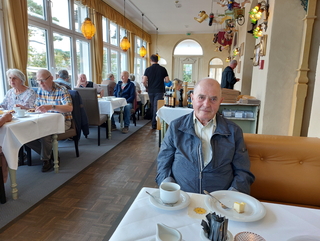 Ein älterer Mann sitzt mit Kaffee und Kuchen an einem Restauranttisch und blickt in die Kamera. Andere Gäste im Hintergrund.