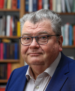 Mann mit grauen Haaren und Brille in blauem Sakko lächelnd, unscharfe Bücherregale im Hintergrund.