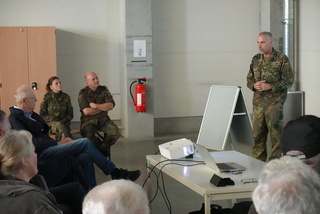 Soldat in Uniform hält Vortrag vor zivilen Zuhörern und zwei Kameraden in einem Raum mit Projektor und Whiteboard.