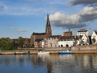 Blick auf eine Stadt am Fluss mit einer großen Kirche, historischen Gebäuden und mehreren Booten unter blauem Himmel mit Wolken.