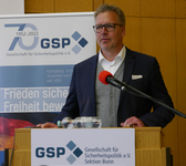 Ein Mann in Sakko und Brille spricht an einem Rednerpult mit GSP-Bannern, die das 70-jährige Jubiläum zeigen.
