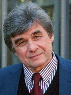 Porträt eines älteren Mannes mit grauem Haar, blauen Augen und leichtem Lächeln, trägt blauen Blazer, gestreiftes Hemd und rote Krawatte.