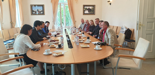 Eine Gruppe von Personen sitzt an einem ovalen Konferenztisch mit Kaffee, Gebäck und Trauben in einem hellen Raum.