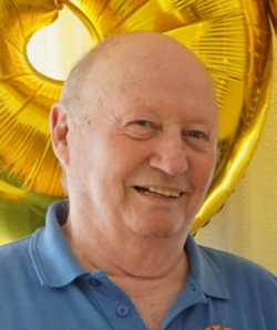 Lächelnder älterer Mann in blauem Poloshirt, mit unscharfen goldenen Feierballons im Hintergrund.