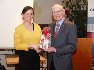 Lächelnde Frau in Gelb und Mann im Anzug schütteln Hände, halten dabei gemeinsam eine Geschenktüte bei einer Veranstaltung.