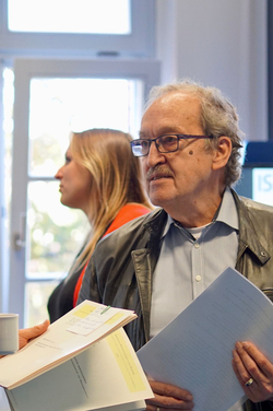 Älterer Mann mit Brille und Schnurrbart hält Dokumente bei einer Veranstaltung. Eine Frau ist unscharf im Hintergrund.