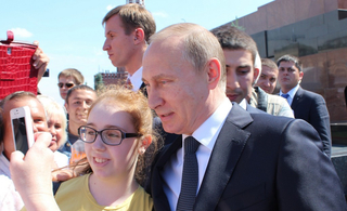 Wladimir Putin steht mit einer jungen Frau, die ein Selfie macht, umgeben von Menschen im Freien.