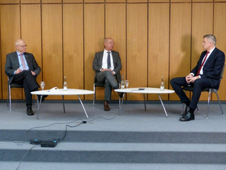 Drei Männer in Anzügen sitzen auf einer Bühne mit kleinen Tischen und Getränken, wahrscheinlich bei einer Diskussion.