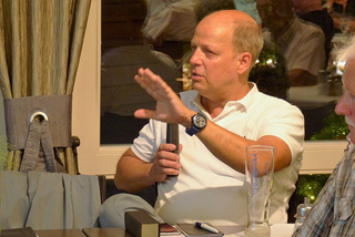 Ein kahler Mann im weißen Poloshirt spricht, mit der rechten Hand gestikulierend und einem Objekt in der linken, Bierglas auf dem Tisch.