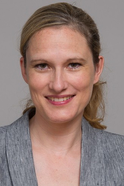 Kopfportrait einer lächelnden Frau mit blonden Haaren, die einen grauen Blazer vor schlichtem Hintergrund trägt.