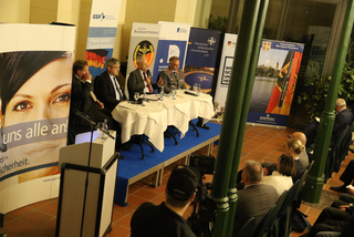 Vier Männer bei einer Podiumsdiskussion vor Bannern von Sicherheits- und Verteidigungsorganisationen, Publikum lauscht.