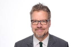 Lächelnder Mann mittleren Alters mit grauem Haar, Brille, Bart, grauem Anzug und grüner Krawatte vor weißem Hintergrund.