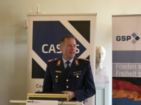 Mann in dunkelblauer Militäruniform spricht am Rednerpult. Im Hintergrund: 'CAS'-, 'GSP'-Banner und eine Deutschlandflagge.