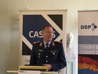 Mann in dunkelblauer Militäruniform spricht am Rednerpult. Im Hintergrund: 'CAS'-, 'GSP'-Banner und eine Deutschlandflagge.