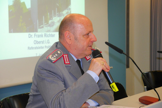 Oberst Dr. Frank Richter in Militäruniform spricht bei einer Präsentation in ein Mikrofon.