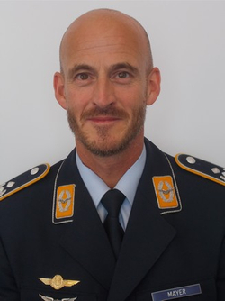 Porträt eines lächelnden, glatzköpfigen Mannes mit Bart, in deutscher Luftwaffe-Uniform mit Namensschild 'MAYER'.