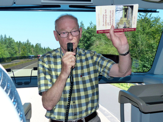 Ein Mann mit Brille, kariertem Hemd, Mikrofon und Geschichtsbuch über eine Synagoge präsentiert auf einer Busfahrt.