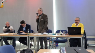 Ein Mann spricht mit Mikrofon an einem Konferenztisch mit drei anderen Männern. Einer schaut auf einen Laptop, andere hören zu.