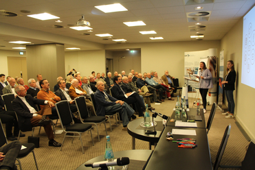Eine Rednerin präsentiert vor einem Publikum von Männern in einem Konferenzraum, mit Social-Media-Symbolen auf einer Leinwand.