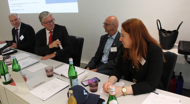 Vier Personen, drei Männer und eine Frau, sitzen an einem Konferenztisch mit Dokumenten, Wasserflaschen und einem Mikrofon.