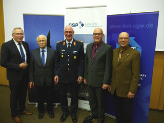 Fünf Männer, vier in Anzügen und einer in Uniform, stehen lächelnd vor Bannern von "Gesellschaft für Sicherheitspolitik e.V." und "dwt-sgw.de".