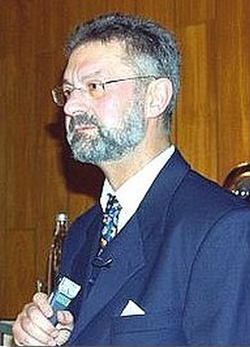 Profil eines bärtigen Mannes mit Brille im dunklen Anzug, weißem Hemd und gemusterter Krawatte, ein kleines Gerät haltend.
