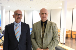 Zwei glatzköpfige Männer in Anzügen stehen nebeneinander. Links mit Brille und Streifenkrawatte, rechts mit gemusterter Krawatte.