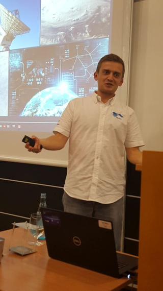 Mann präsentiert Technologiethemen mit Fernbedienung, vor einem Bildschirm mit Satelliten-, Erd- und Netzwerkbildern.