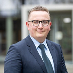 Porträt eines lächelnden Mannes mit Brille, blauem Anzug und Krawatte vor unscharfem Hintergrund.