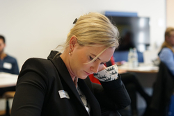 Blonde Frau mit Namensschild, in schwarzem Blazer und gemustertem Hemd, konzentriert nach unten blickend bei einem professionellen Meeting.