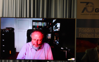 Bärtiger Mann mit Brille und Headset auf einem Bildschirm während eines Videocalls. Daneben ein Banner der Gesellschaft für Sicherheit.