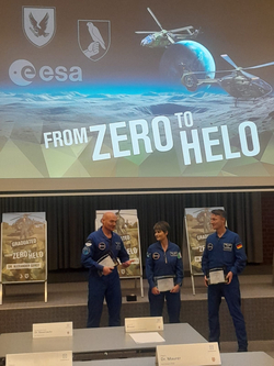 Drei Personen in Fliegeranzügen erhalten Auszeichnungen bei einer ESA-Veranstaltung vor einem Bildschirm mit 'FROM ZERO TO HELO'.