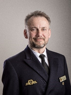 Porträt eines lächelnden Mannes in dunkler Uniform mit Bart und einer goldenen Anstecknadel.