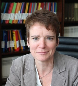 Professionelle Frau mit kurzen braunen Haaren und ernstem Ausdruck, im Sakko, vor einem Bücherregal.
