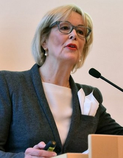 Blonde Frau mit Brille spricht an einem Rednerpult mit Mikrofon und hält einen Presenter.