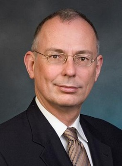 Porträt eines Mannes mit Brille, dunklem Anzug und gestreifter Krawatte, leicht lächelnd vor einem melierten dunklen Hintergrund.